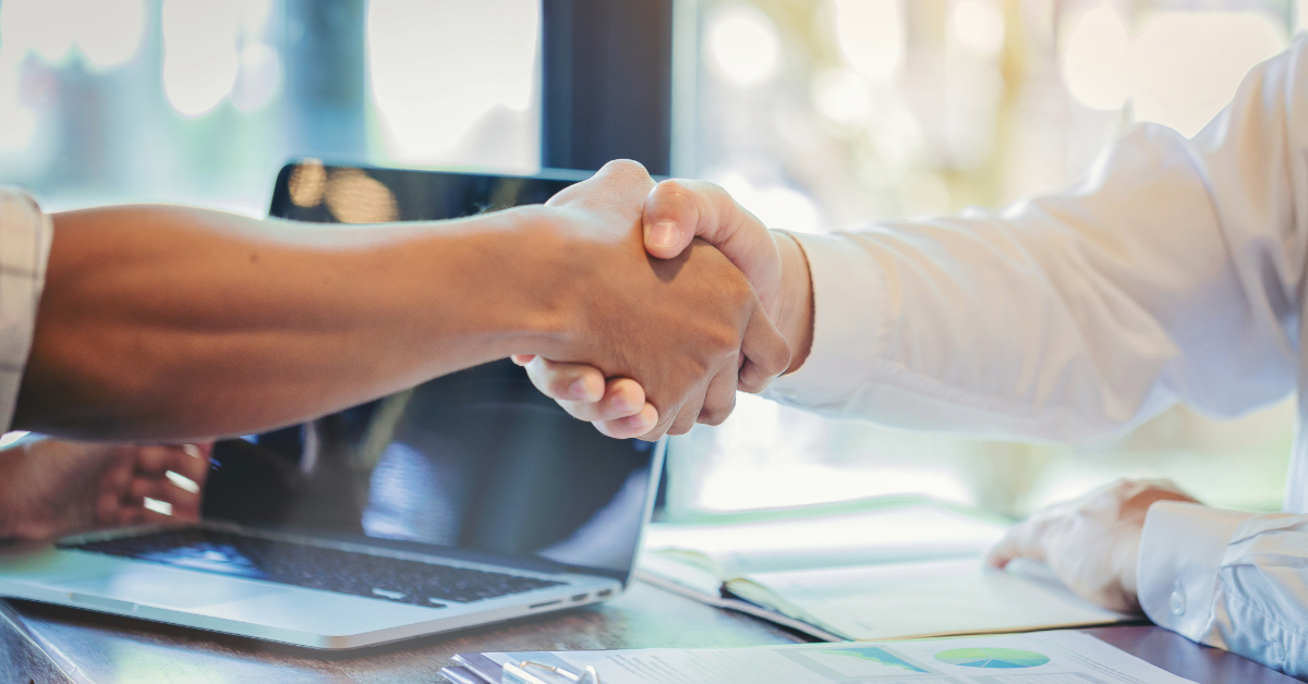 Two individuals shaking hands over a table, exploring business loan opportunities, with documents and a laptop in the background.