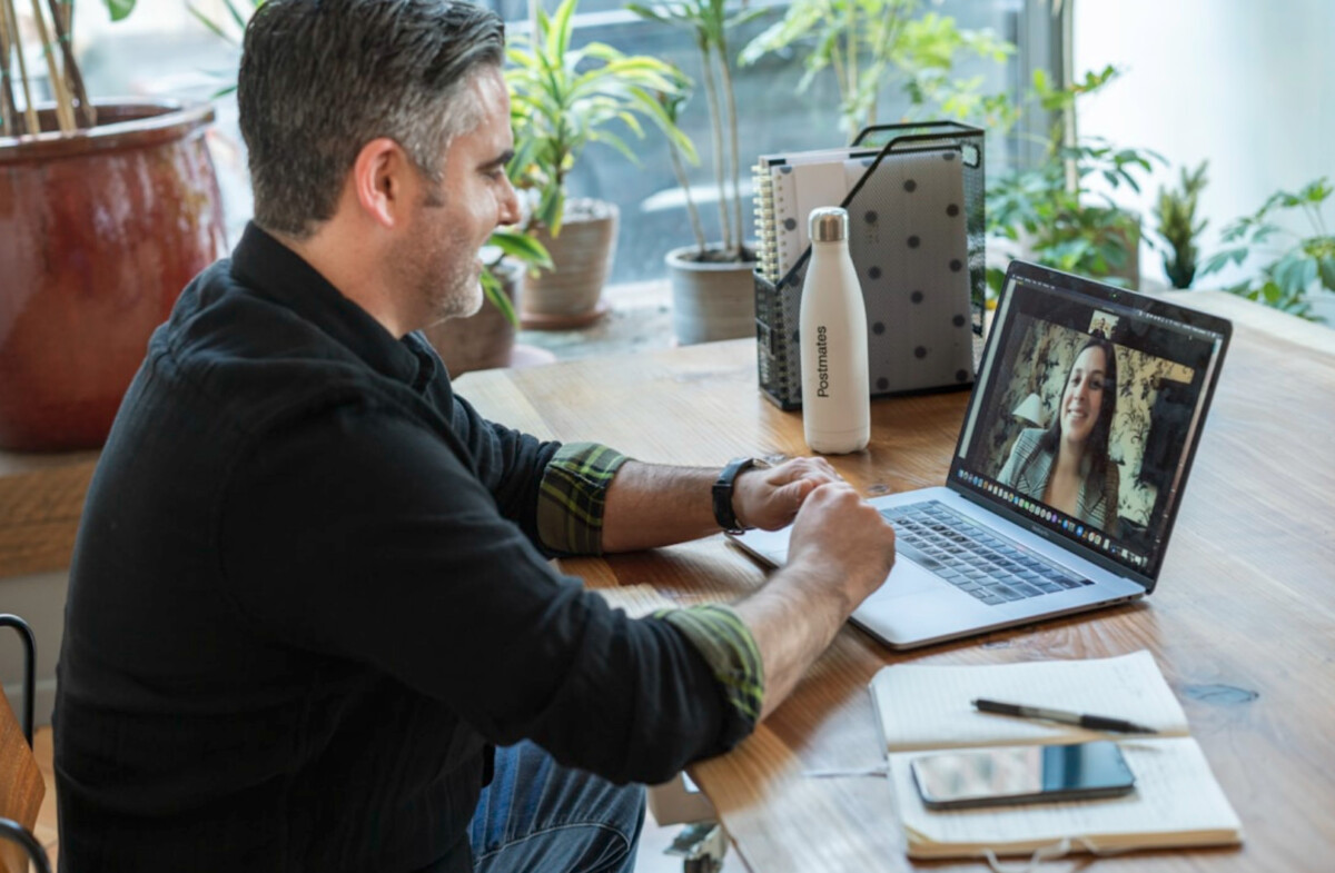 A man is sitting at a desk, video conferencing on a laptop with a woman about sales strategies. A notebook, pen, and water bottle lie nearby as plants add a serene touch to the background.