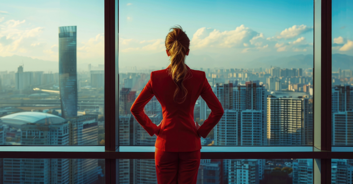 A person in a red suit stands with hands on hips, gazing out at a city skyline through a large window, envisioning innovative solutions and leading companies towards team success during the daytime.