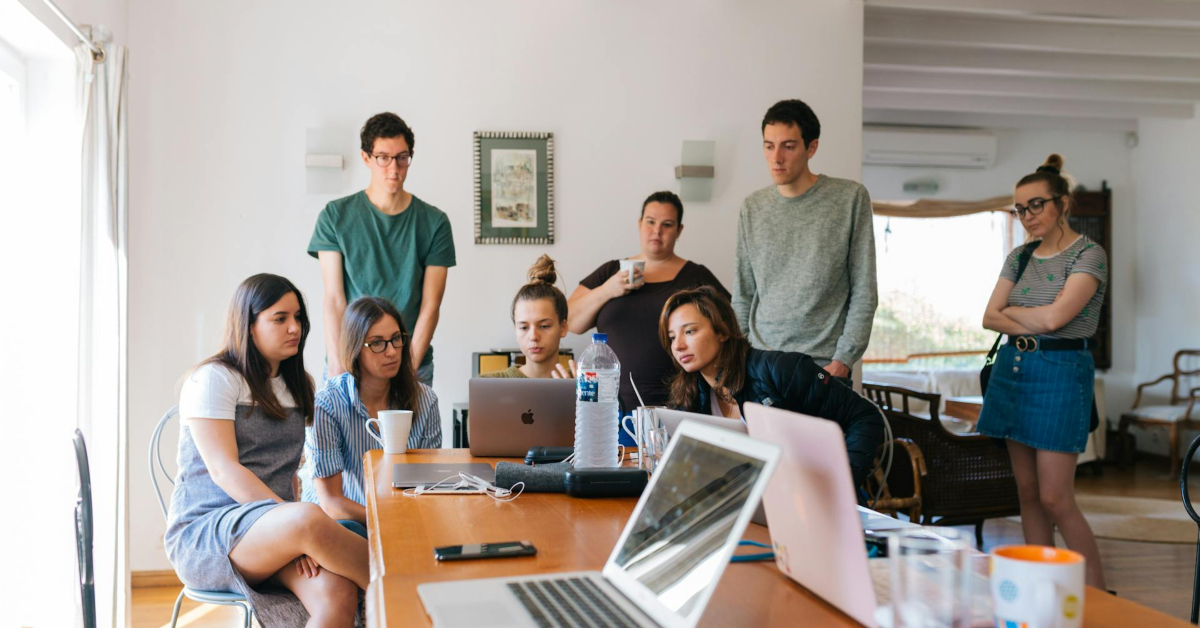 A group of employees gather around a table with multiple laptops, appearing focused and engaged in discussion. The well-lit room, filled with natural light from a nearby window, creates an atmosphere that empowers them to explore effective ways to collaborate.
