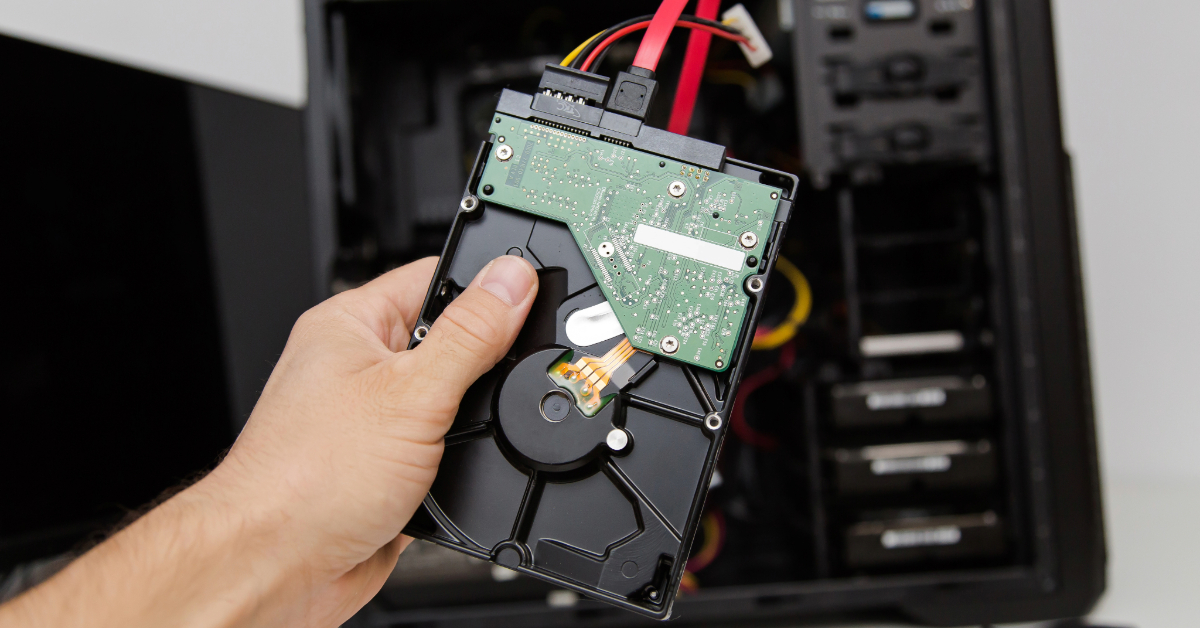 A hand carefully holds a computer hard drive with attached cables in front of an open computer case, ready to safely clone the HDD.
