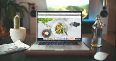 A laptop displaying a salad recipe website sits on a desk with a cactus, bottled water, and a newspaper. Large speakers and a blurred screen loom in the background, creating an inspiring setup to build your ideal business website.
