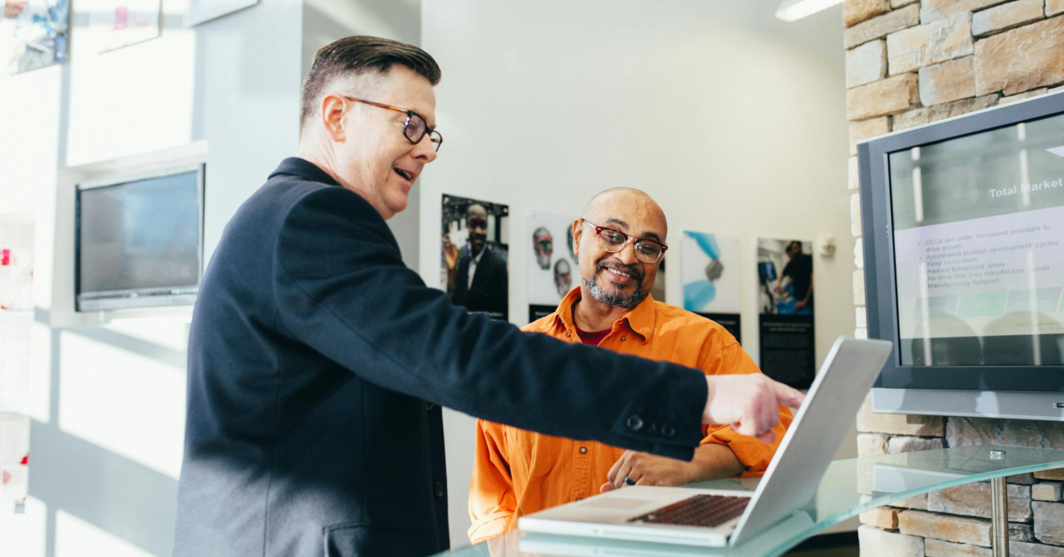 Two men are standing at a counter with an open laptop. One man is pointing at the screen, highlighting their market competence, and both are smiling.