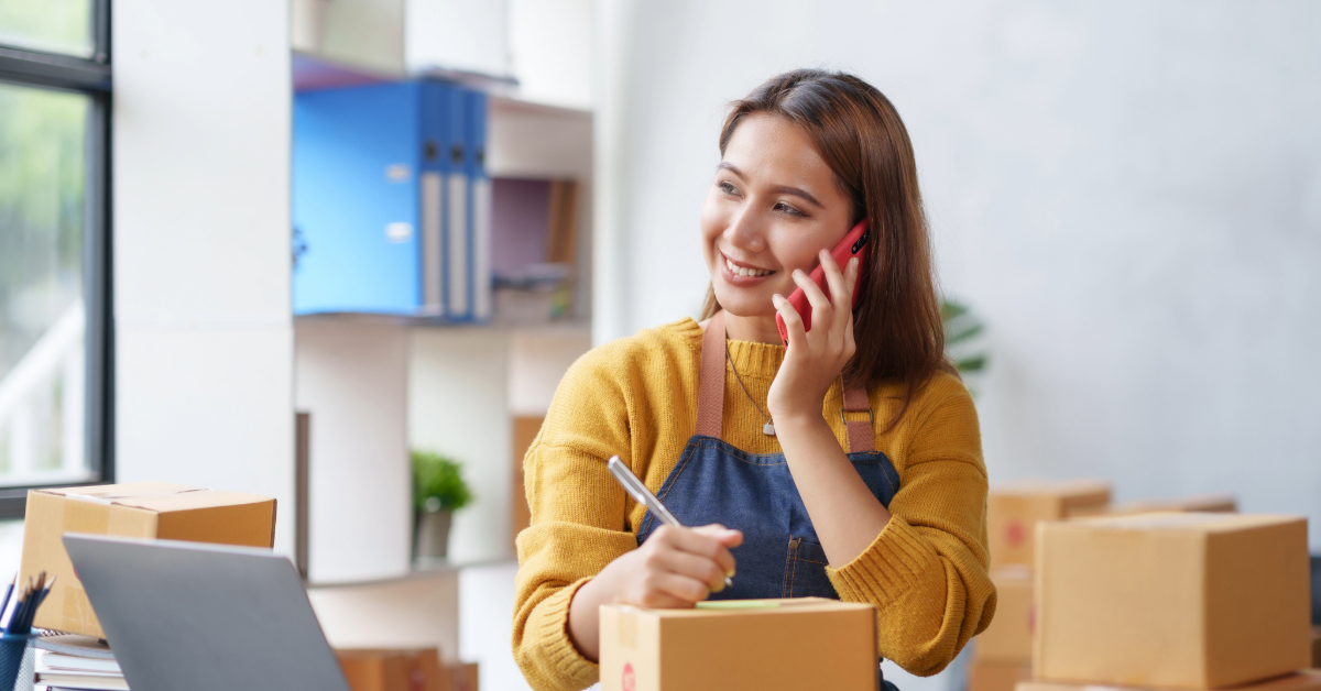 In a bustling mixed-use office, a woman in a yellow sweater and apron juggles e-commerce duties. Surrounded by strategic placements of cardboard boxes and her trusty laptop, she chats on the phone while jotting down notes with precision and flair.