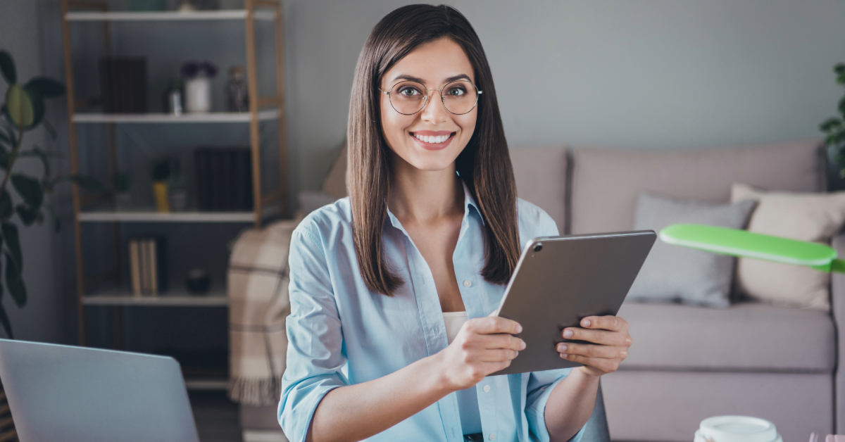 A woman with glasses holding a tablet sits at a desk in a room with shelves and a couch, embodying the essence of entrepreneurs by effortlessly streamlining tasks like a skilled virtual assistant.