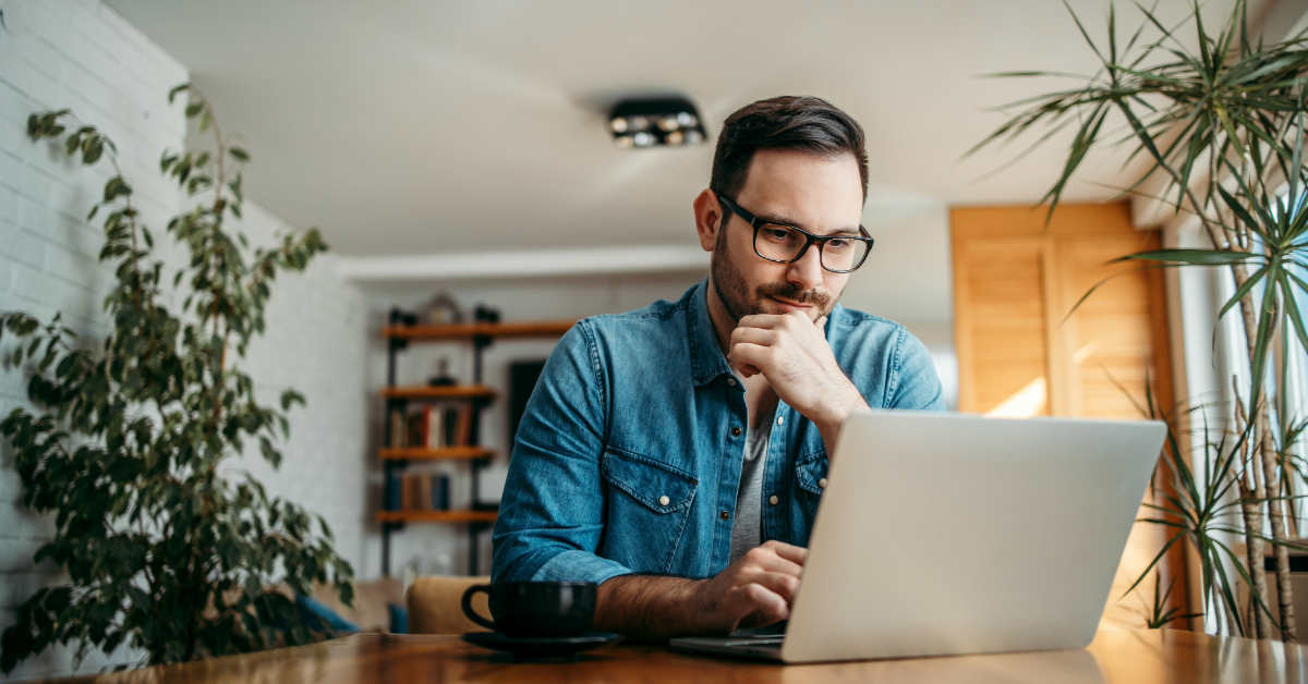 A man with glasses in a denim shirt, sits at a wooden table, exploring options on his laptop. He's making a smart choice over coffee; plants and a bookshelf are nearby as he seeks the ideal liquidity partner.