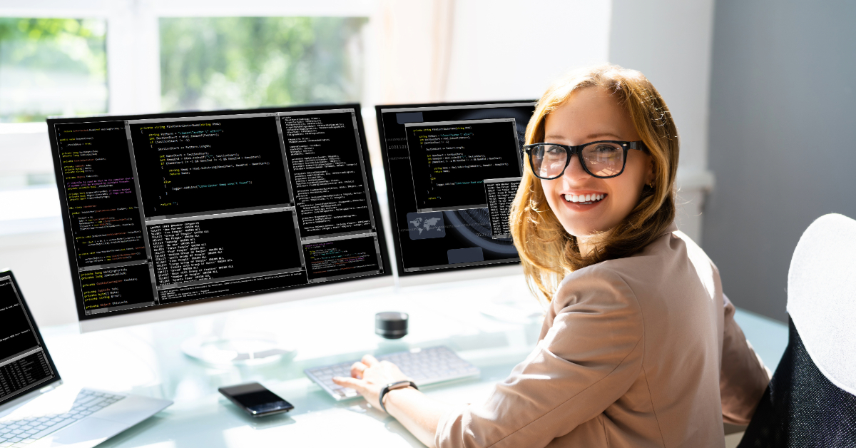 A woman wearing glasses is smiling while sitting at a desk with multiple computer monitors displaying code, reflecting the skills overview of successful C# developers in high demand.