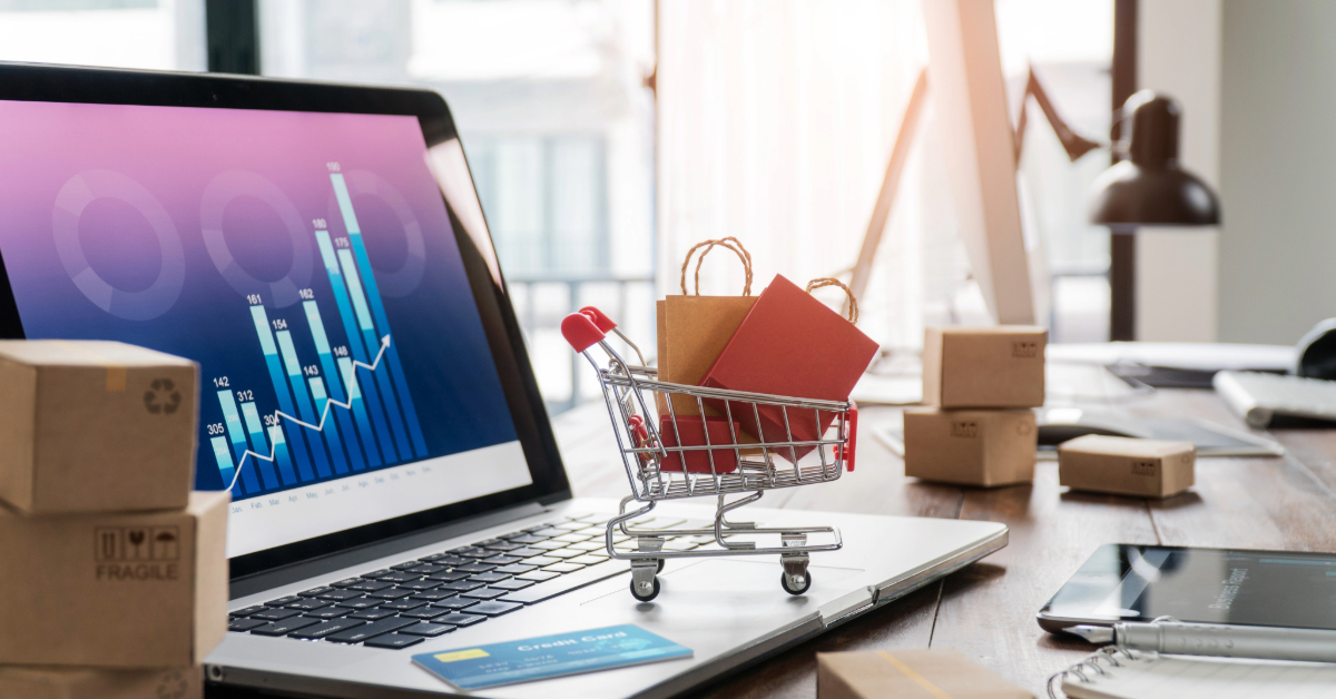 A laptop displaying graphs sits beside a miniature shopping cart holding bags and a credit card, embodying an eCommerce guide on what you need to know. Nearby, small boxes rest on the desk, while a smartphone and lamp complete the backdrop.