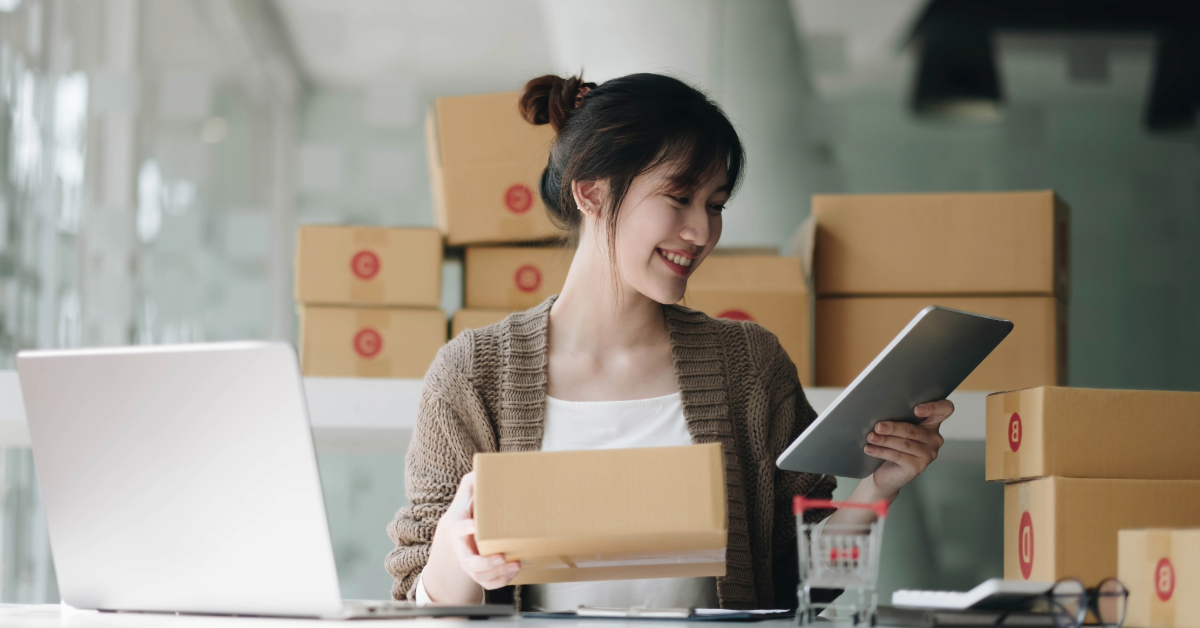 A successful woman sits at her desk, juggling a tablet and a cardboard box, a scene characteristic of 2024's e-commerce boom. Behind her, multiple stacked boxes hint at a bustling venture in progress.