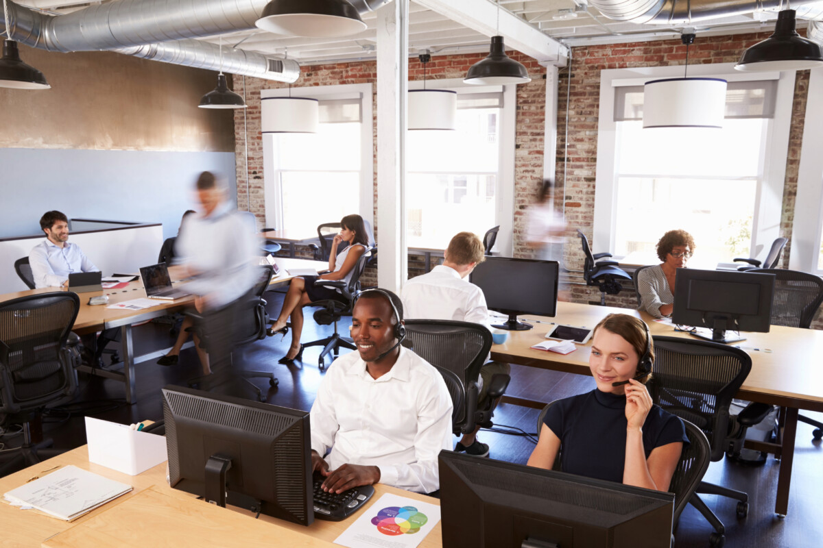Modern open-plan office bustling with diverse employees engaged in BPO solutions, working on computers and talking on phones. The space features exposed brick walls and large windows, creating an inspiring environment for ecommerce outsourcing and customer support excellence.
