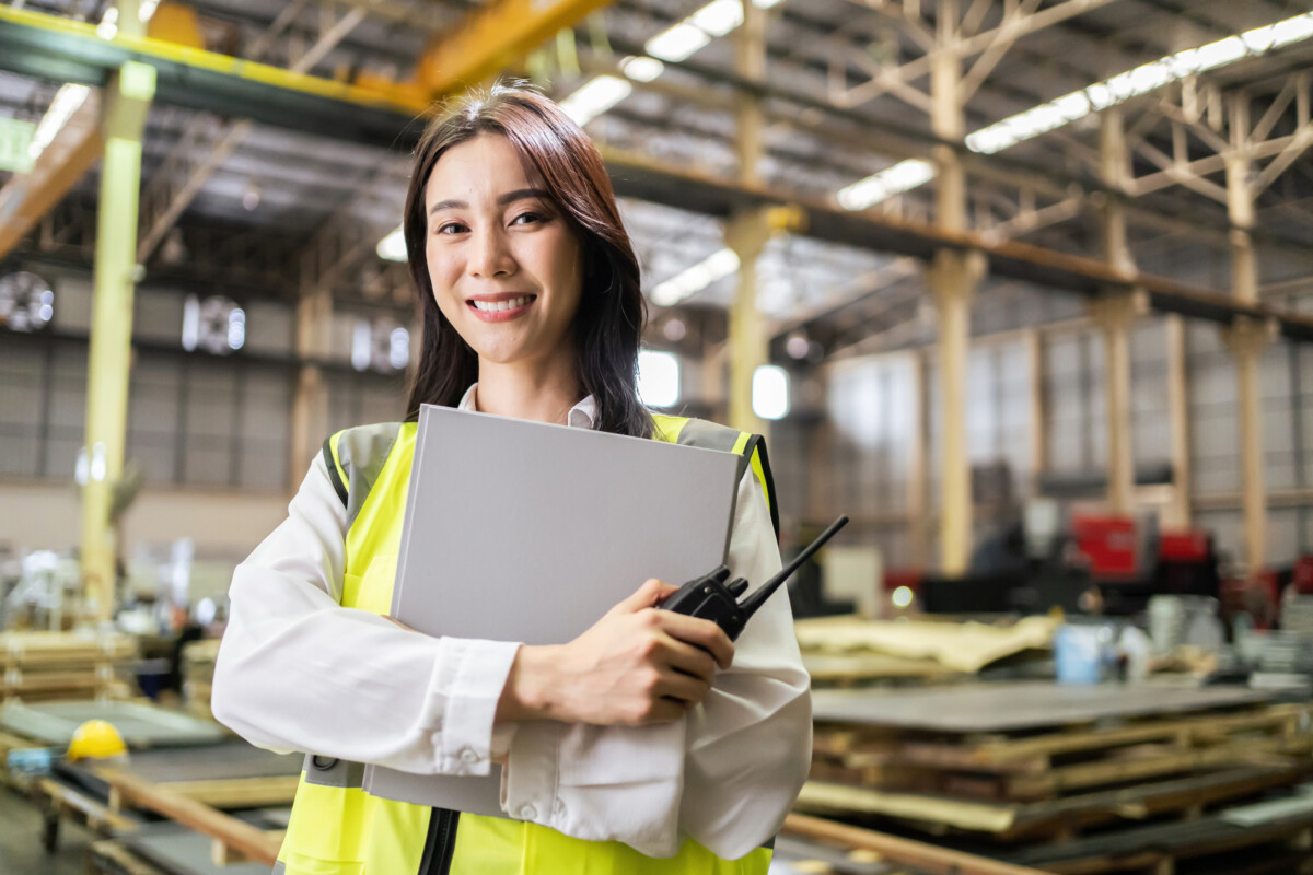 In a spacious warehouse, a person in a high-visibility vest holds a clipboard and radio, expertly coordinating activities. With the right product from reliable manufacturers stacked around them, they ensure efficiency and precision.