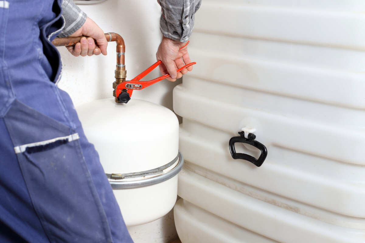 A professional in work attire uses a wrench to adjust a valve on a white cylindrical tank beside a larger plastic tank, showcasing expertise in plumbing services.