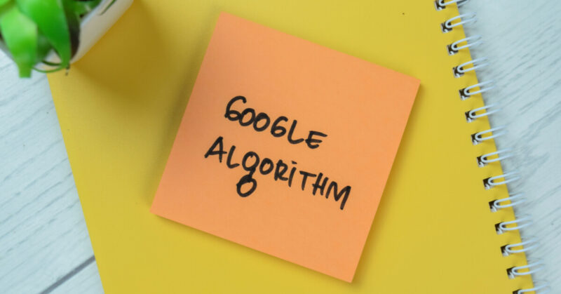 A yellow sticky note with "Google Algorithm" written on it rests atop a yellow notebook, hinting at SEO strategies for E-Commerce Sites. Nearby, a small green plant adds a touch of nature to the workspace focused on mastering the Google March 2024 Core Update.
