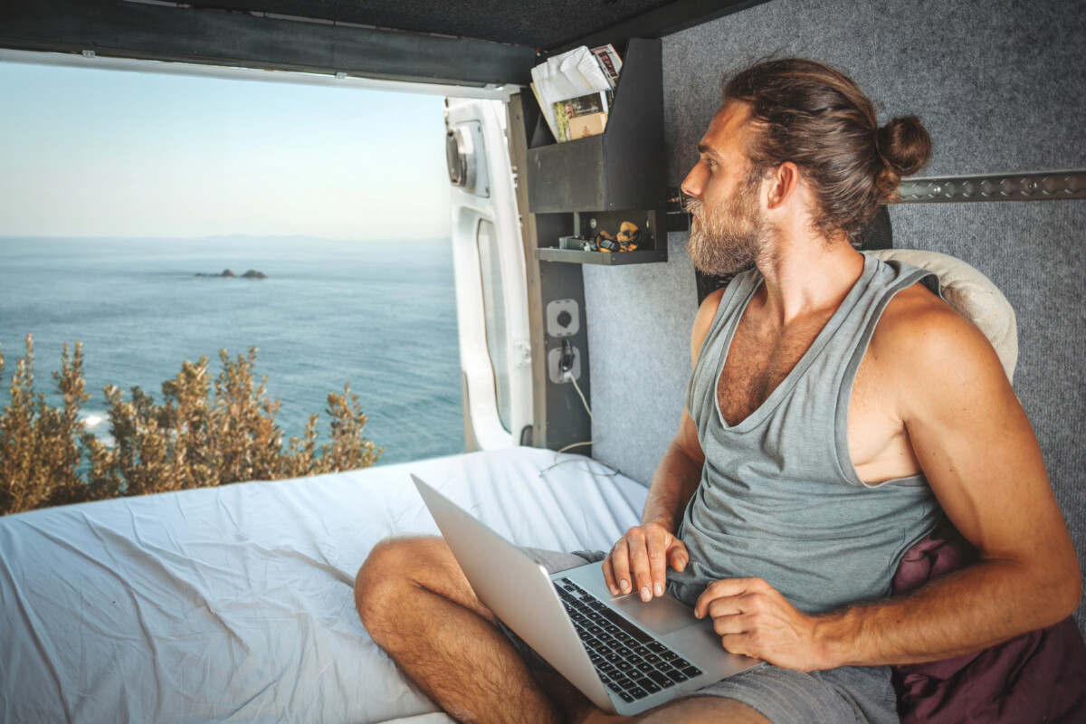 Man with a laptop sits in a van, embracing digital nomadism as he gazes out at the ocean through an open door, balancing work and leisure perfectly.