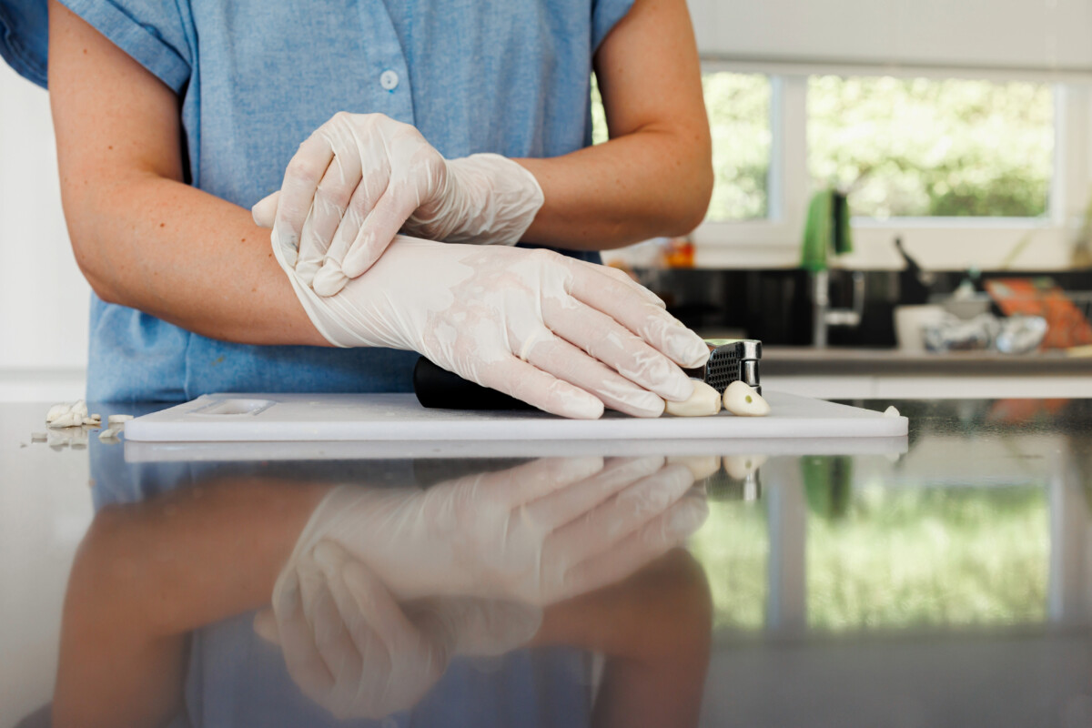 In a kitchen that values health and safety standards, a person wearing gloves carefully slices garlic on a cutting board.