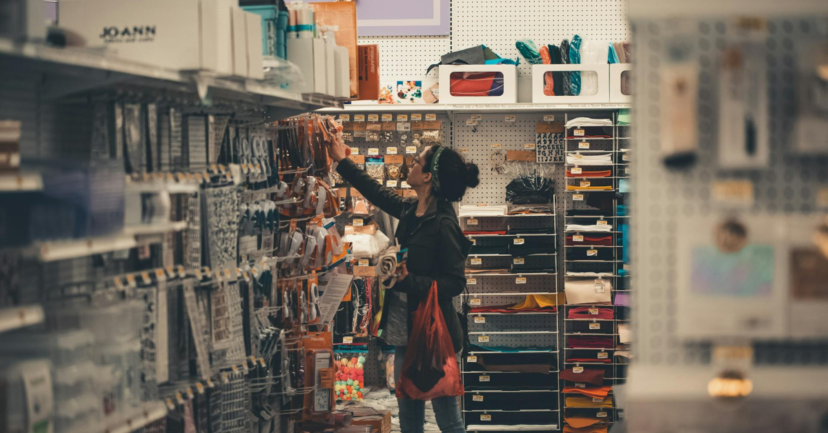A customer enjoys their shopping experience as they reach for an item on the shelf, holding a red tote bag. The store aisle is lined with shelves filled with various products and supplies.