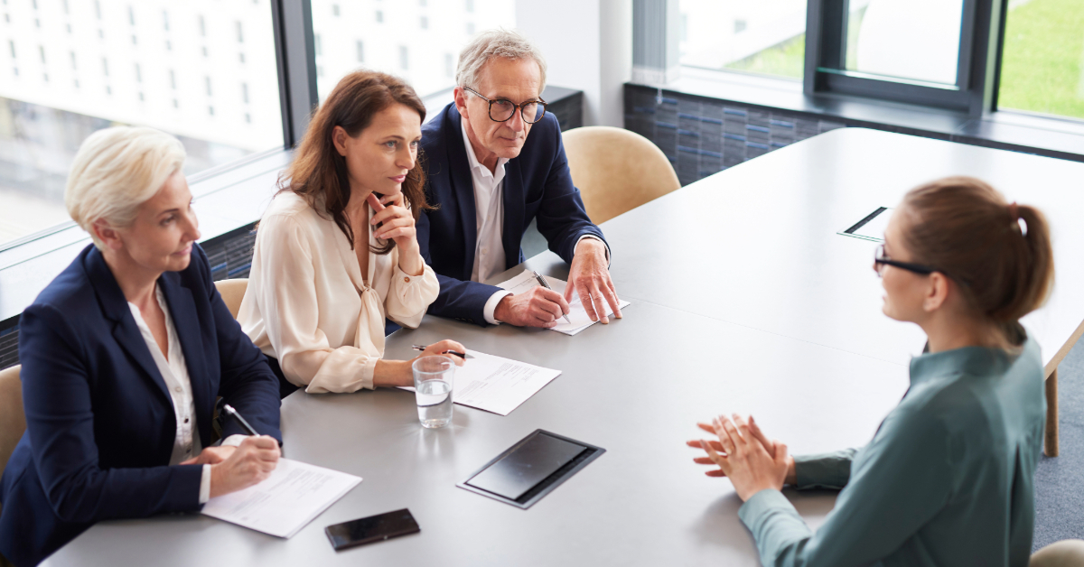 A person sits across from three individuals conducting an interview in a modern office setting, employing a stellar marketer recruitment strategy to ensure they find the perfect candidate.