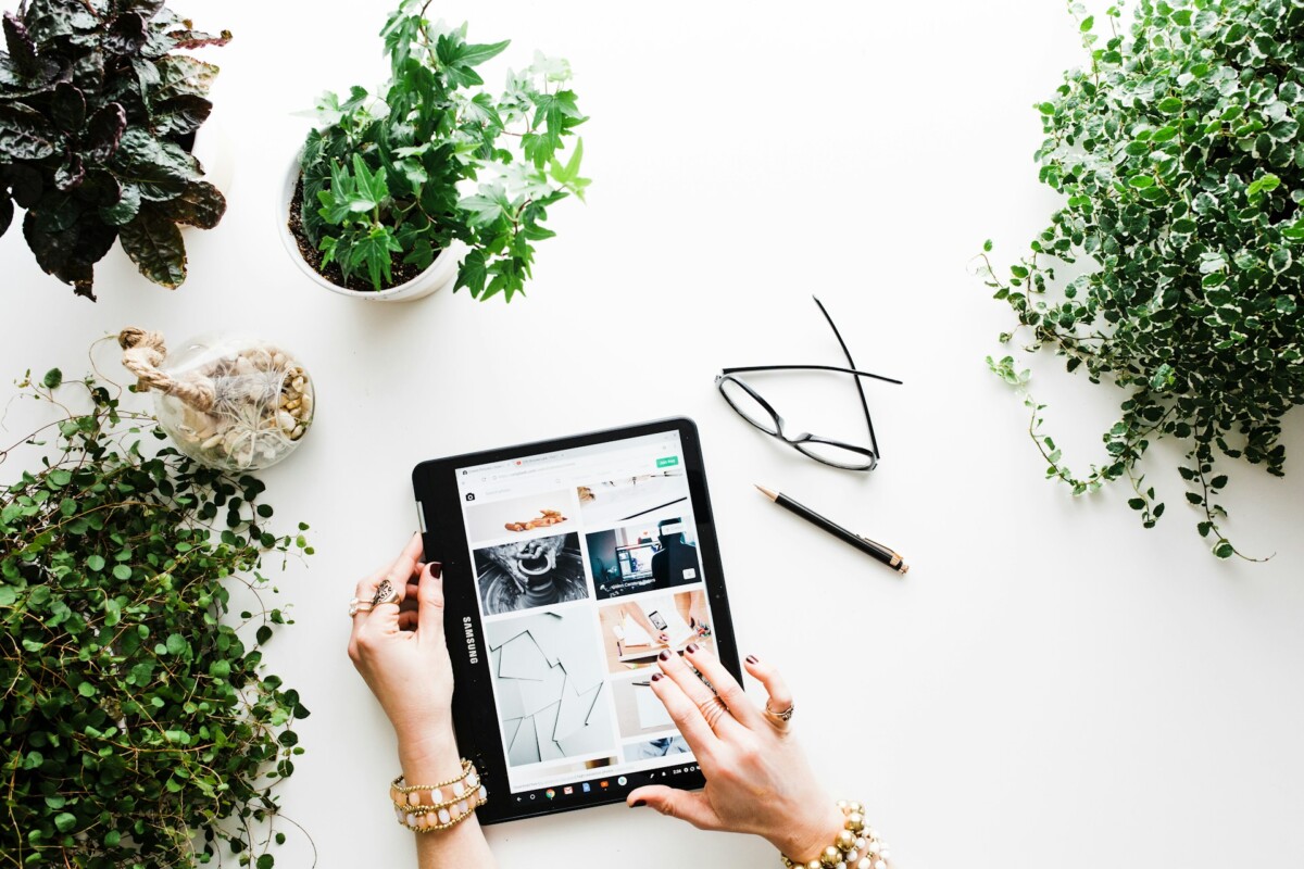 Hands hold a tablet on a white table surrounded by potted plants, a pair of glasses, and a pen, demonstrating how integrating ecommerce gamification can boost engagement in everyday activities.