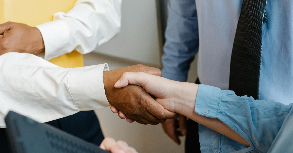 Two people are shaking hands in an office setting, surrounded by colleagues. One is wearing a white shirt, and the other a light blue one, marking the beginning of a promising business partnership.
