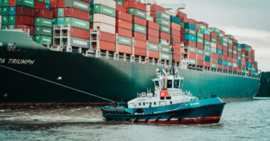 A small blue tugboat skillfully navigates the water, guiding a large cargo ship loaded with multicolored shipping containers, illustrating essential transformations in the supply chain.