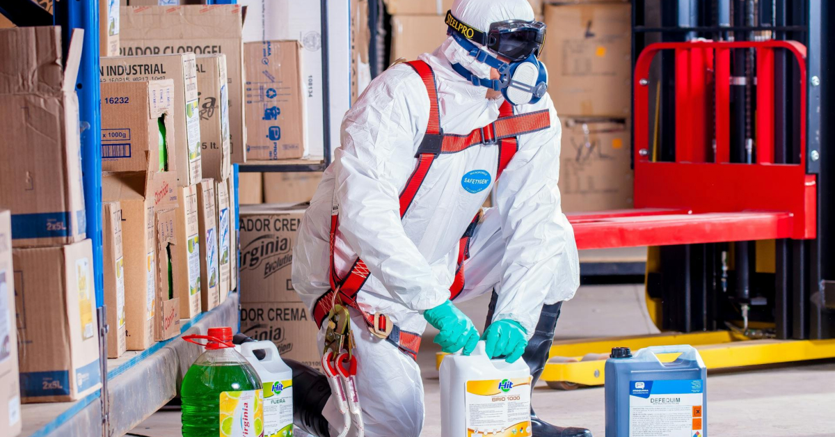 A person in protective gear is meticulously handling toxic substances in a storage area, surrounded by boxes with a forklift nearby, exemplifying excellent hazard management and workplace safety practices.