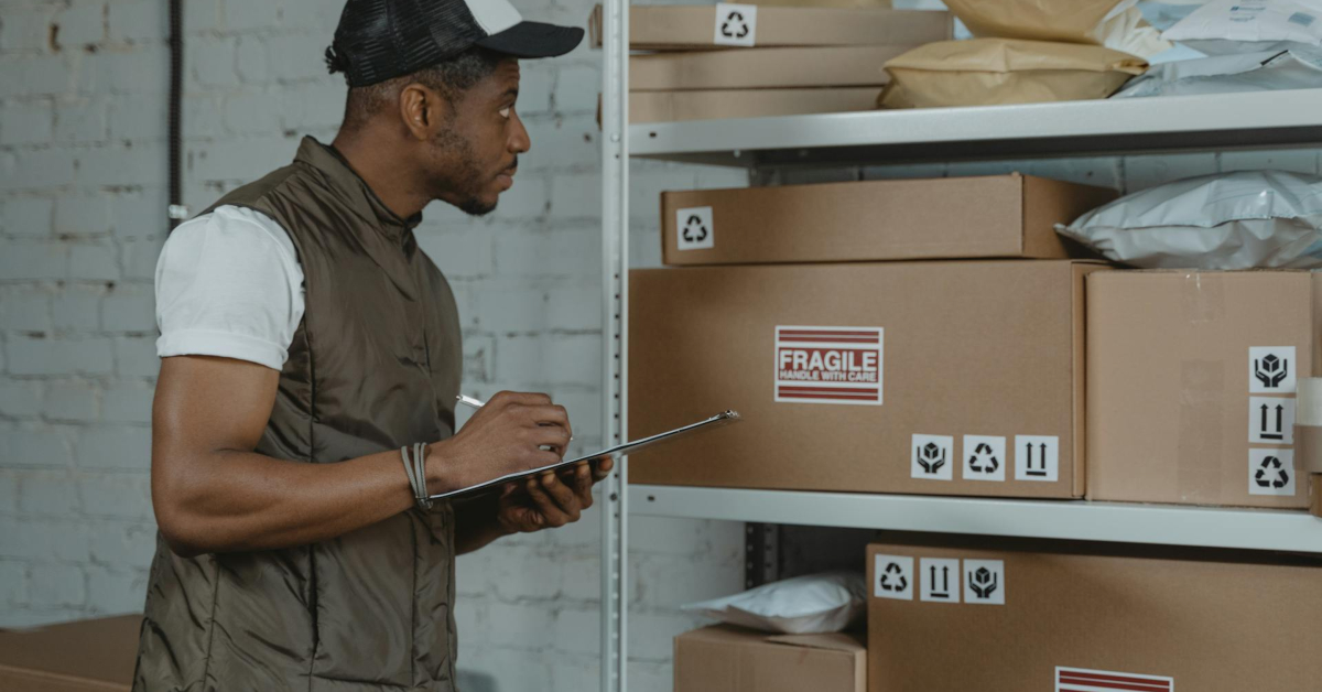 A person in a cap and vest holding a clipboard hums softly while examining shelves filled with various labeled packages and boxes, expertly managing the logistics of the bustling storage area.