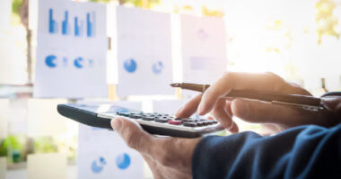 A person uses a calculator and holds a pen, analyzing charts and graphs that reveal the true costs behind common myths of running a business.
