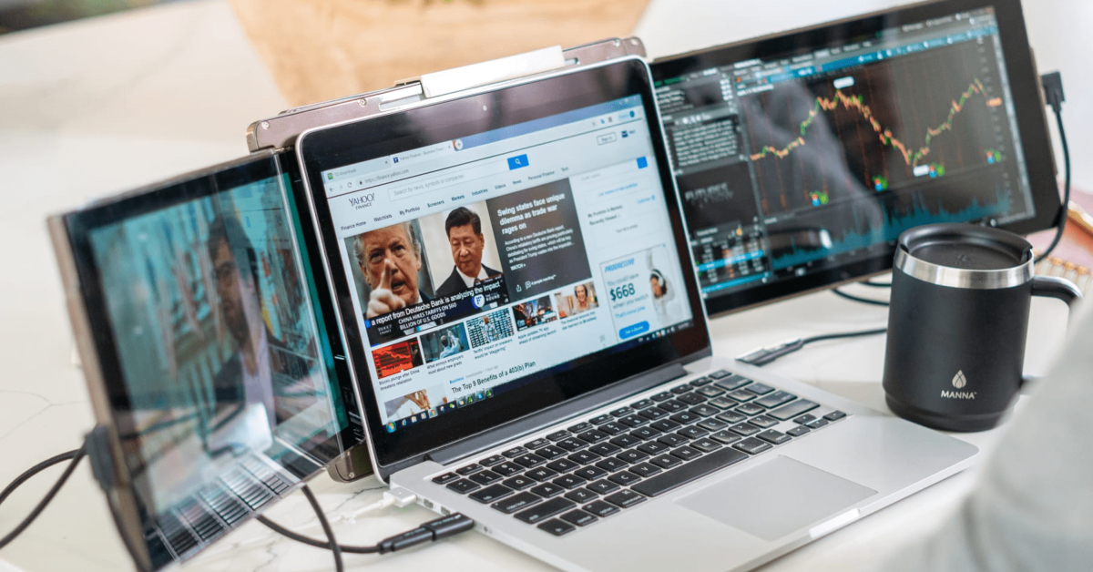 A laptop with financial news on the screen is surrounded by two eco-friendly portable monitors displaying stock market charts. A black insulated cup sits nearby, embodying a seamless blend of technology and sustainability.