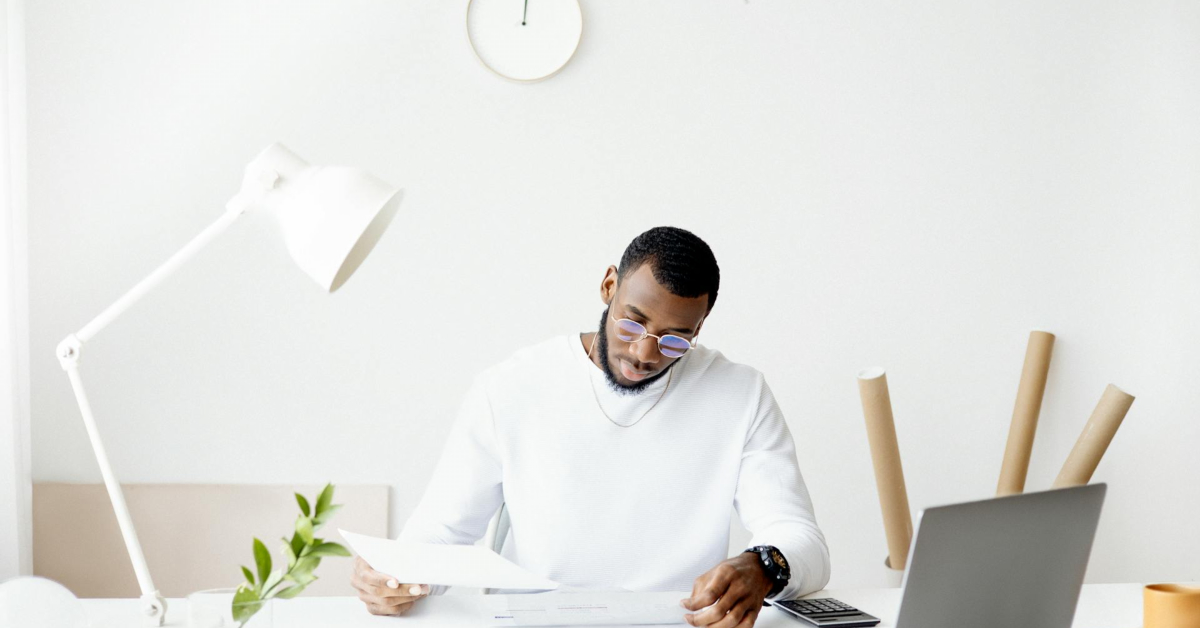 A man in a white sweater sits at a desk in his spare room, examining papers on his laptop. A white lamp and potted plant add charm to the workspace, while three cardboard tubes hint at small business ambitions breaking out of the box.