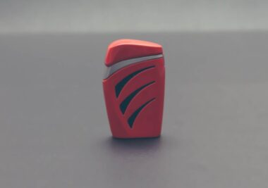 A red and black, curved rectangular lighter with sleek design patterns stands on a gray surface, sparking inspiration for the next steps in product idea development.