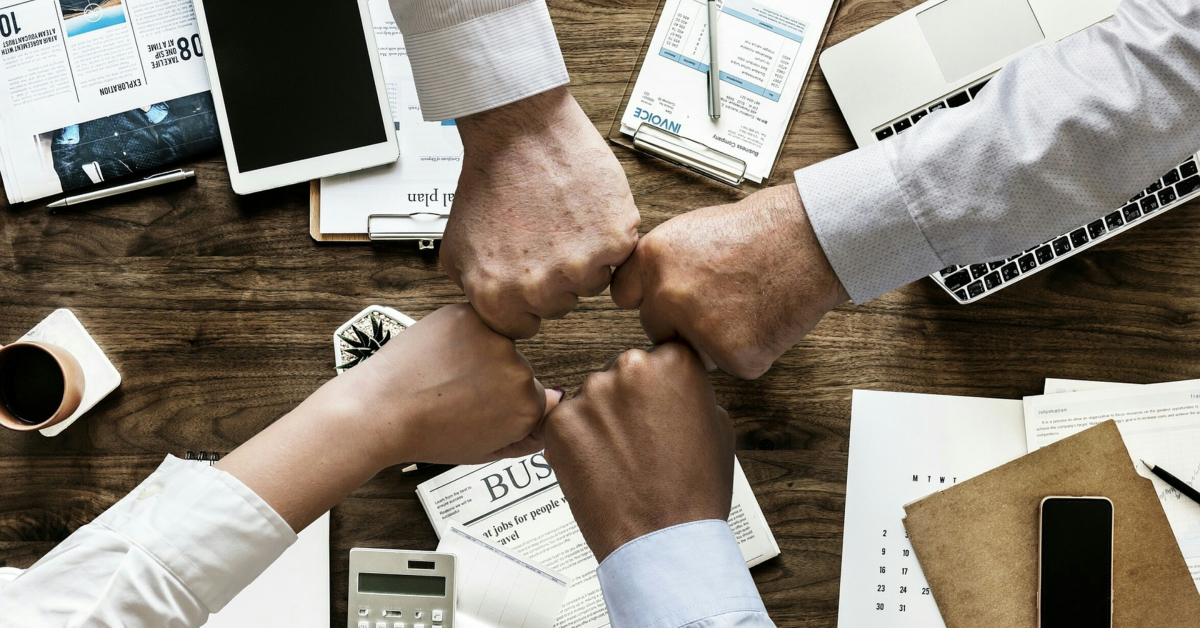 Four hands fist-bumping over a wooden table adorned with newspapers, a laptop, a tablet, and coffee—a symbol of small businesses mastering budget management.