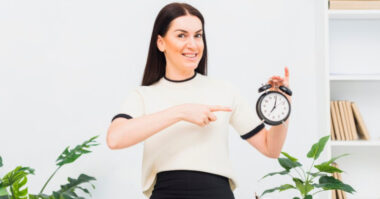 A woman in a white shirt smiles and points at a black alarm clock, emphasizing the importance of time management, standing next to green plants against a white background.