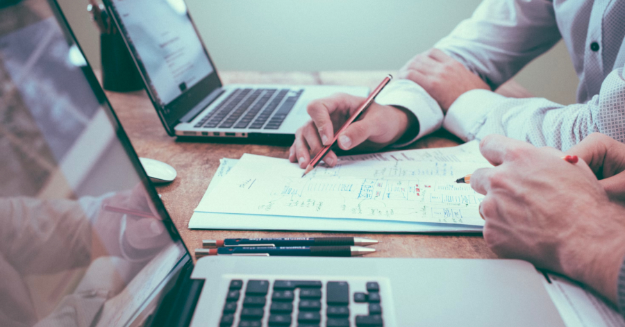 Two people collaborating at a table with laptops and documents discuss inventory costs. One points at a sheet of paper with a pencil, highlighting the economic order quantity, while the other listens attentively.
