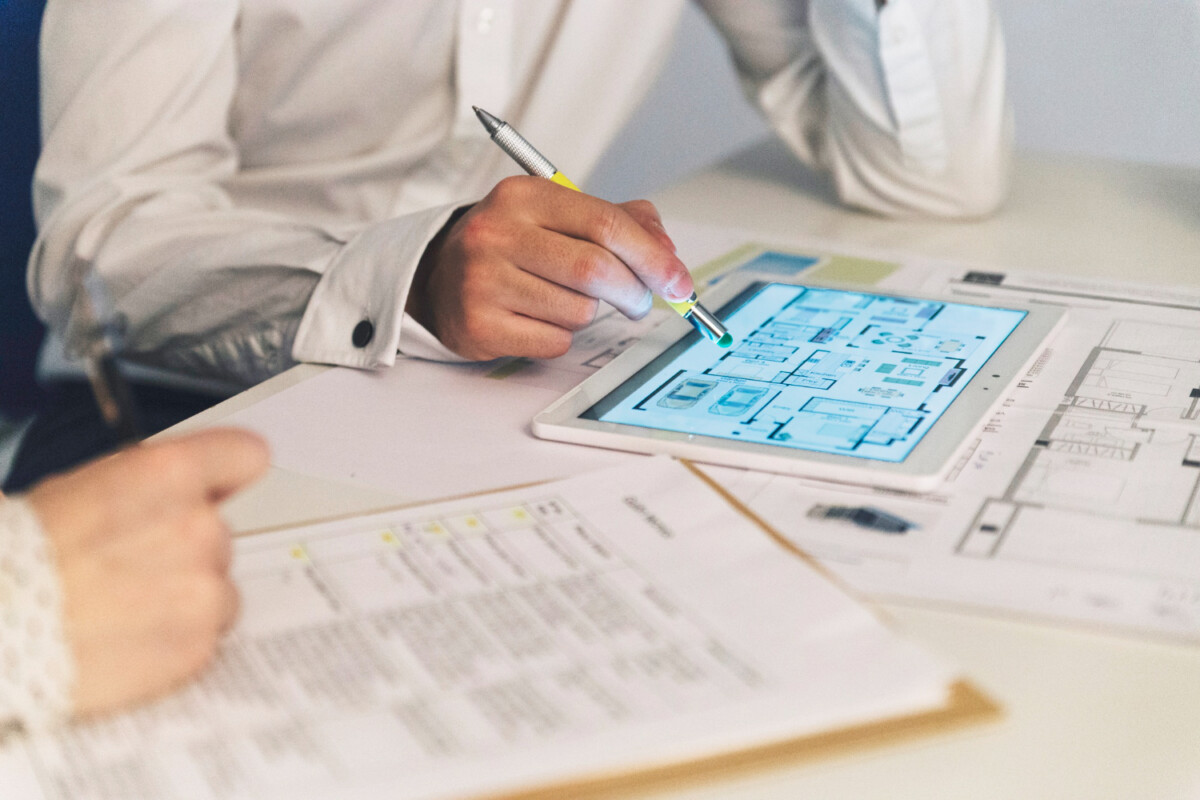 Two people review high-quality architectural blueprints on a tablet and paper documents, discussing plans at a desk with a pen and clipboard, surrounded by detailed technical illustrations.