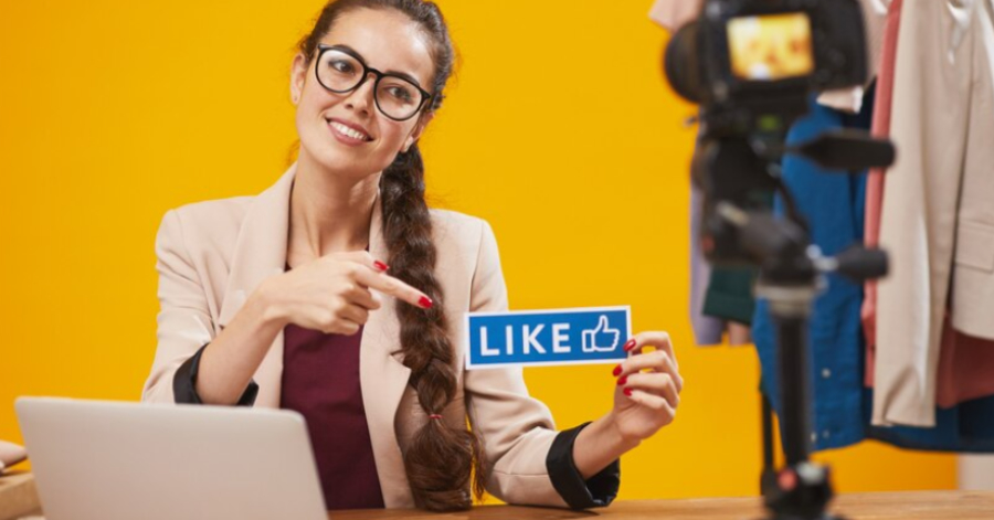Woman with glasses sits at a desk with a laptop, pointing to a "LIKE" sign while being filmed by a camera—demonstrating live stream promotion strategies to help in boosting viewership, with a yellow background and clothes rack visible.
