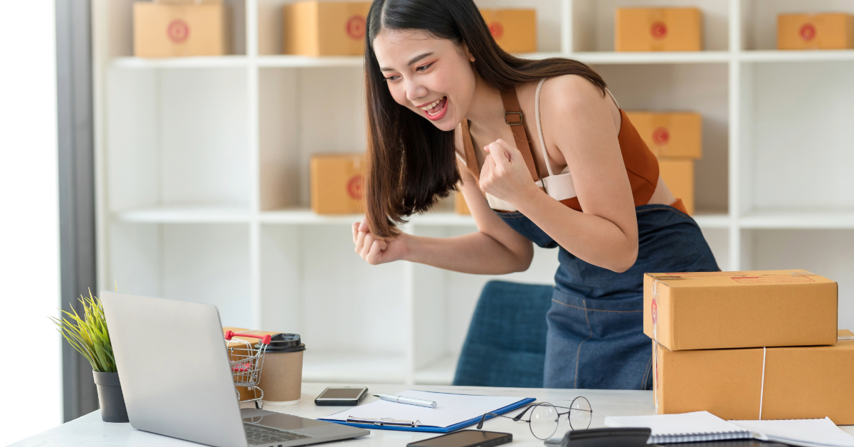 A woman in an apron stands at a desk with packages, looking excitedly at a laptop, embracing the online transition to e-commerce in a workspace with shelves filled with boxes.