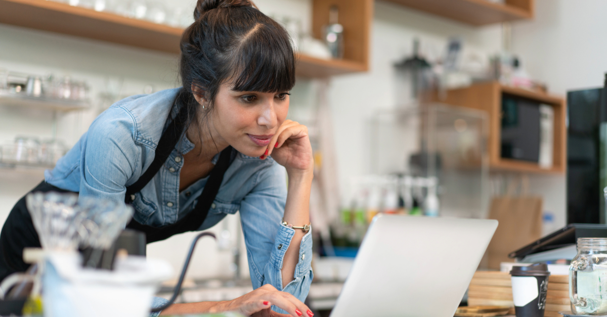 Woman wearing an apron stands at a counter, leaning forward and looking at a laptop screen in a modern, well-lit café—perhaps researching how to start an online coffee business.