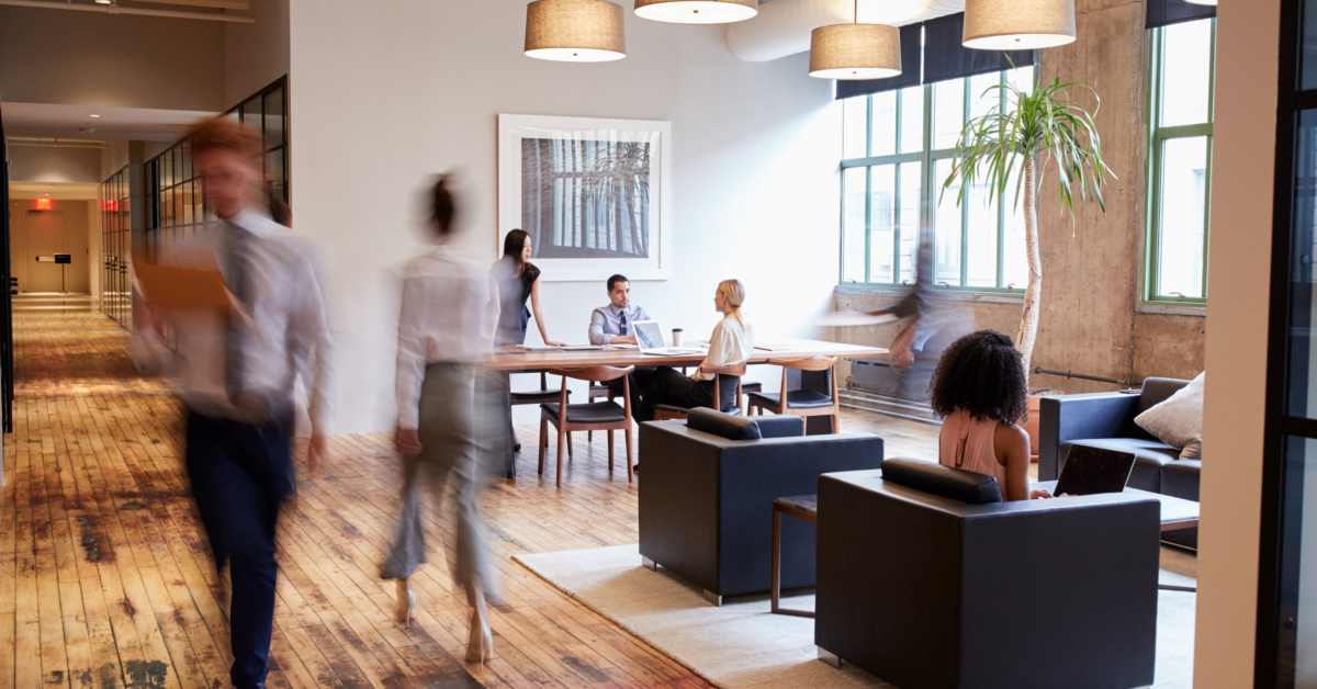 Modern open office space with people working at tables and sitting in armchairs; some individuals are walking through the wooden-floored ultimate office, filled with large windows and indoor plants.