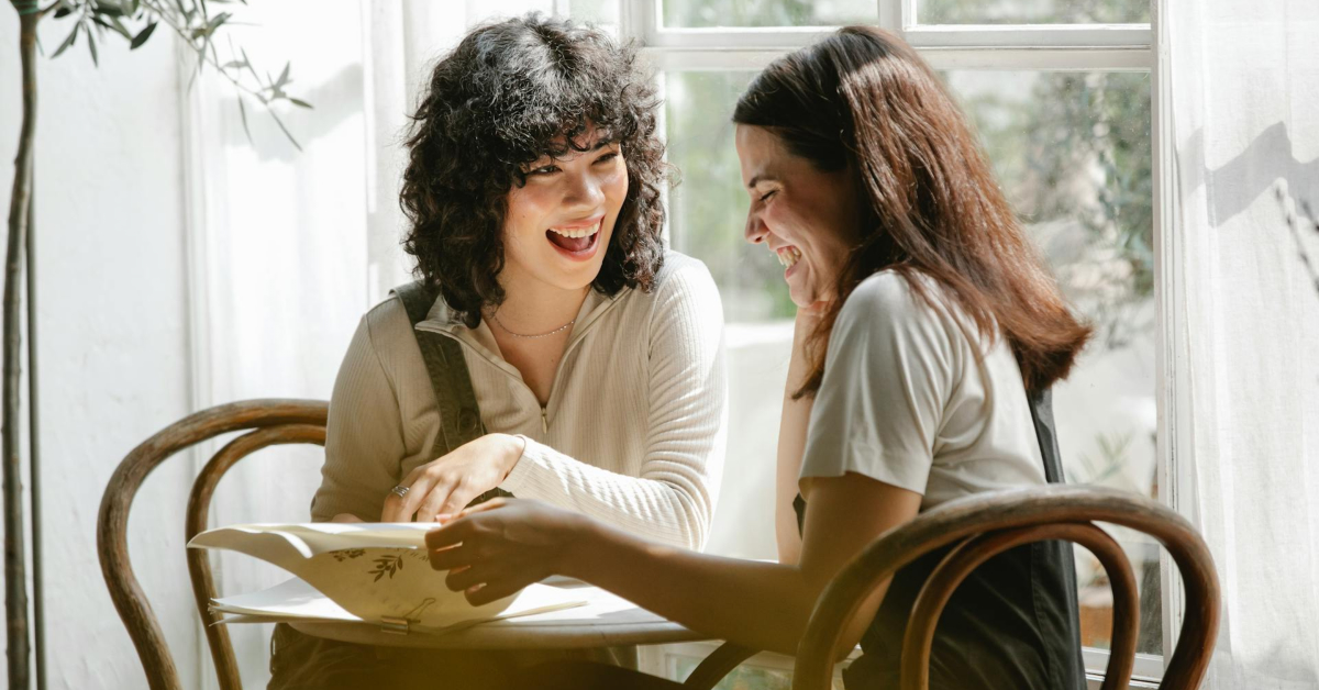 Two women sit at a table by a window, smiling and looking at an open book together in a bright room, reflecting workplace wellbeing and the importance of supporting employees.