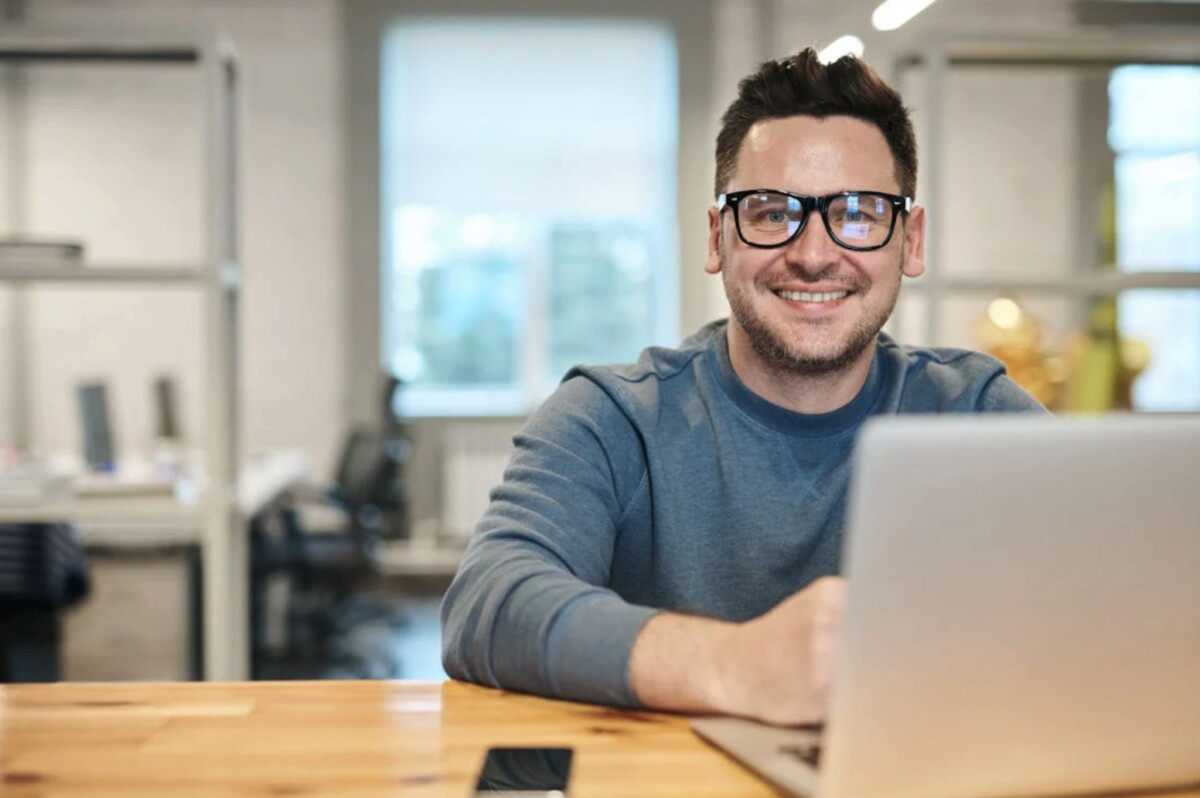 A man wearing glasses and a blue sweater sits at a desk, smiling at the camera, with a laptop and smartphone in front of him in a modern office—ready to manage his ecommerce business on Shopify.