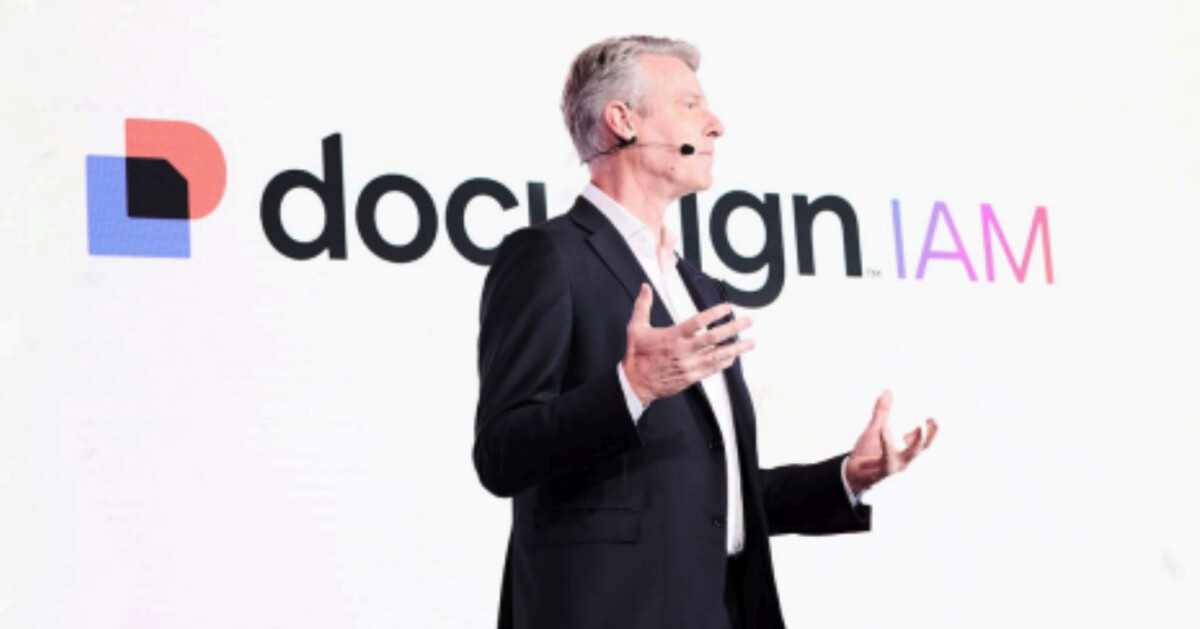 A man in a suit speaks on stage with a headset microphone in front of a screen displaying the DocuSign IAM logo, capturing the energy of event photography.