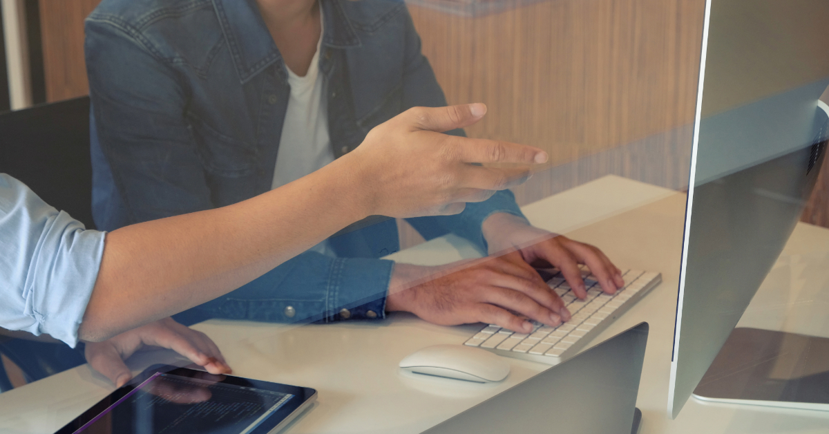 Two people sit at a desk; one is typing on a keyboard while the other gestures toward a computer screen. A tablet with code and dynamic test cases highlights their focus on software excellence.