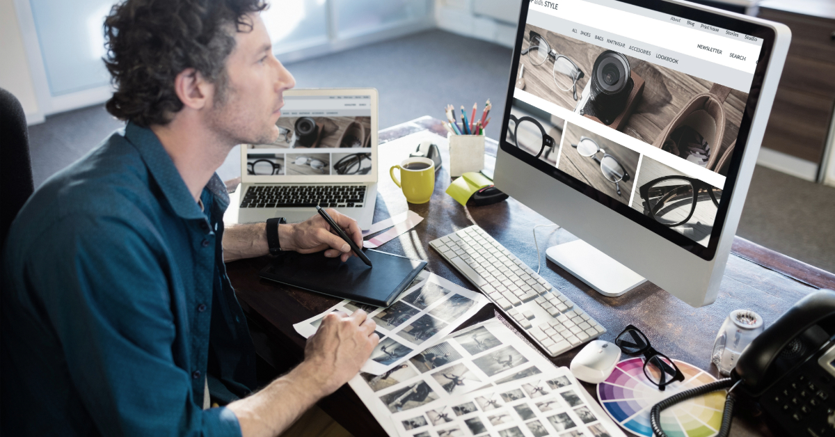 Man working at a desk with a computer, laptop, drawing tablet, and photo prints, editing images on the screen in a modern office with a user-friendly interface designed for driving sales.