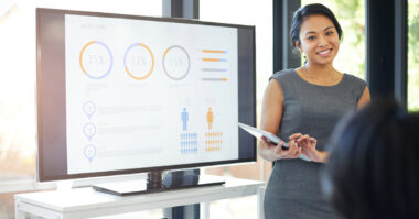A woman in business attire stands next to a screen displaying charts and graphs, holding a tablet and demonstrating effective presentations to an audience in a bright office.
