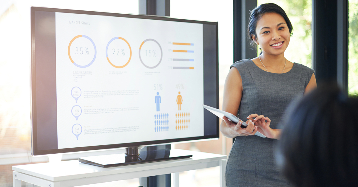 A woman in business attire stands next to a screen displaying charts and graphs, holding a tablet and demonstrating effective presentations to an audience in a bright office.
