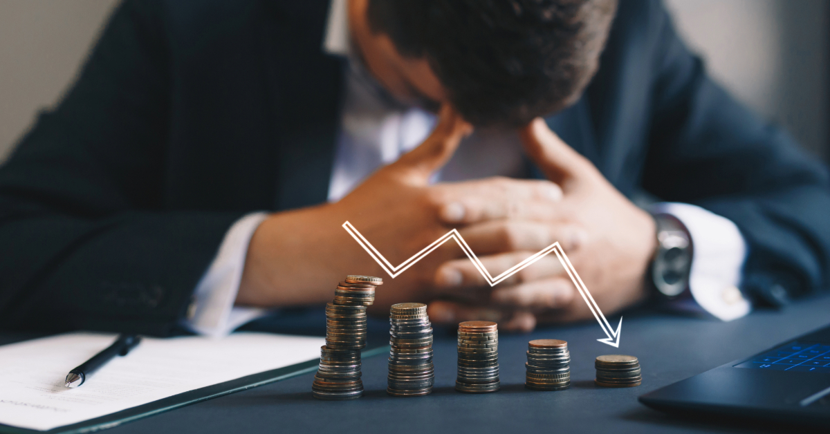 A person in a business suit sits with head bowed behind stacked coins and a downward trending arrow, highlighting financial loss and the importance of budgeting to avoid bankruptcy.