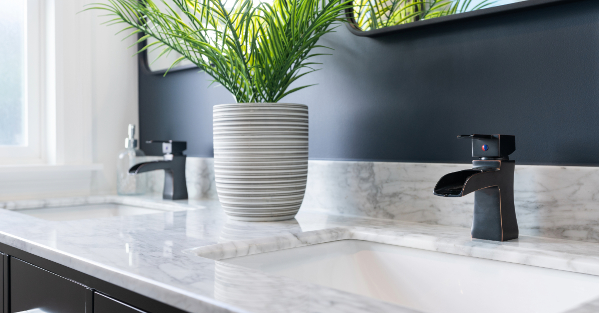 A modern bathroom countertop with two sinks, stylish black bathroom fixtures, a potted plant in a striped vase, and a dark wall with a mirror—perfectly showcasing today’s trending bathroom accessories.