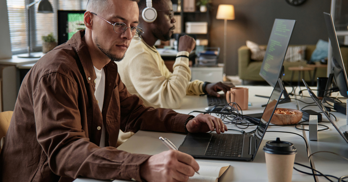 Two men work at computers in a modern office. One writes in a notebook while looking at his laptop, studying an Appium guide; the other, wearing headphones, types at his desktop. Coffee cups are on the desk.