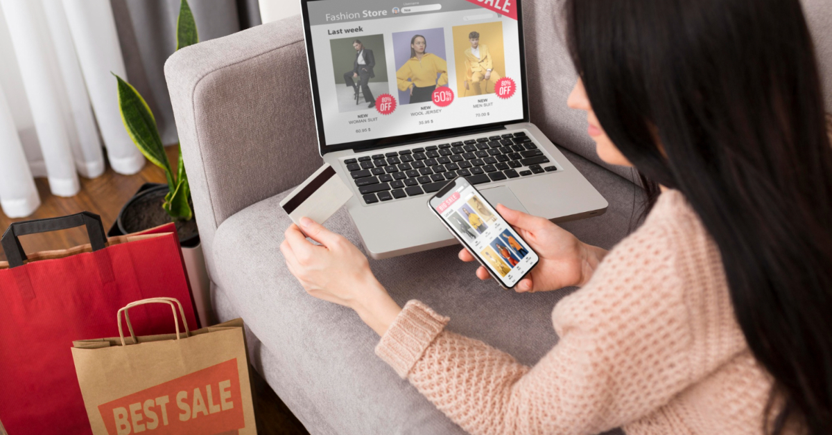 Woman sitting on a couch shops online using her laptop and smartphone, holding a credit card; shopping bags labeled "Best Sale" are on the floor, highlighting the impact of E-Commerce Marketing.