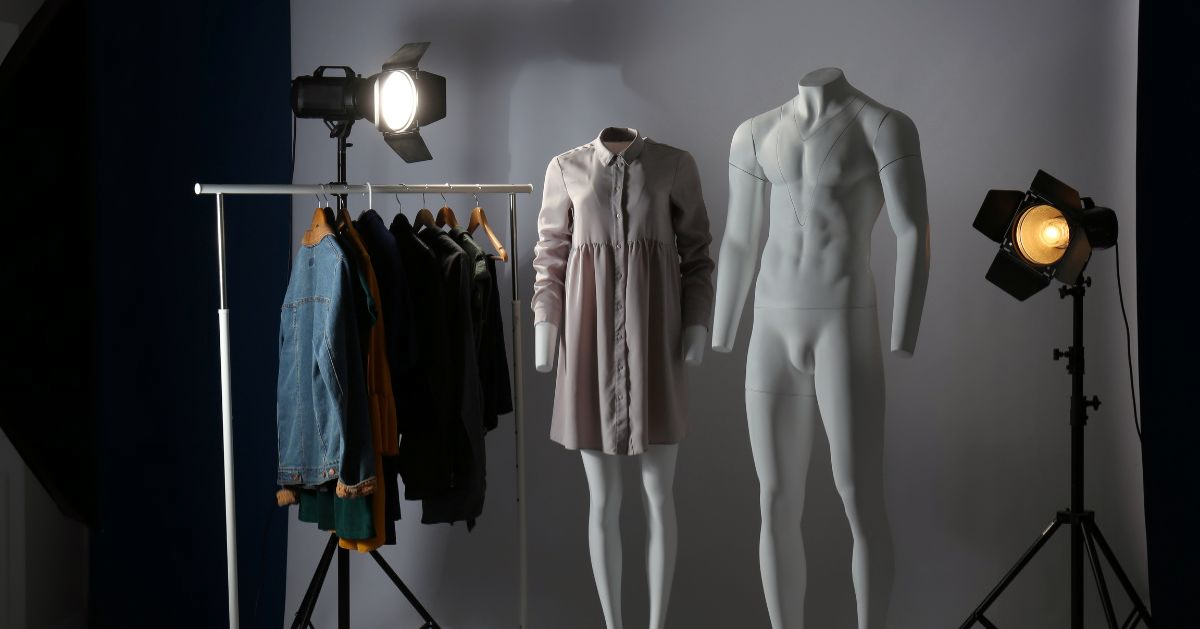 Two white mannequins stand under studio lights—one in a light dress, the other unclothed. A clothing rack with various garments is on the left, showcasing how a Ghost Mannequin Service boosts any online clothing business's visual appeal.
